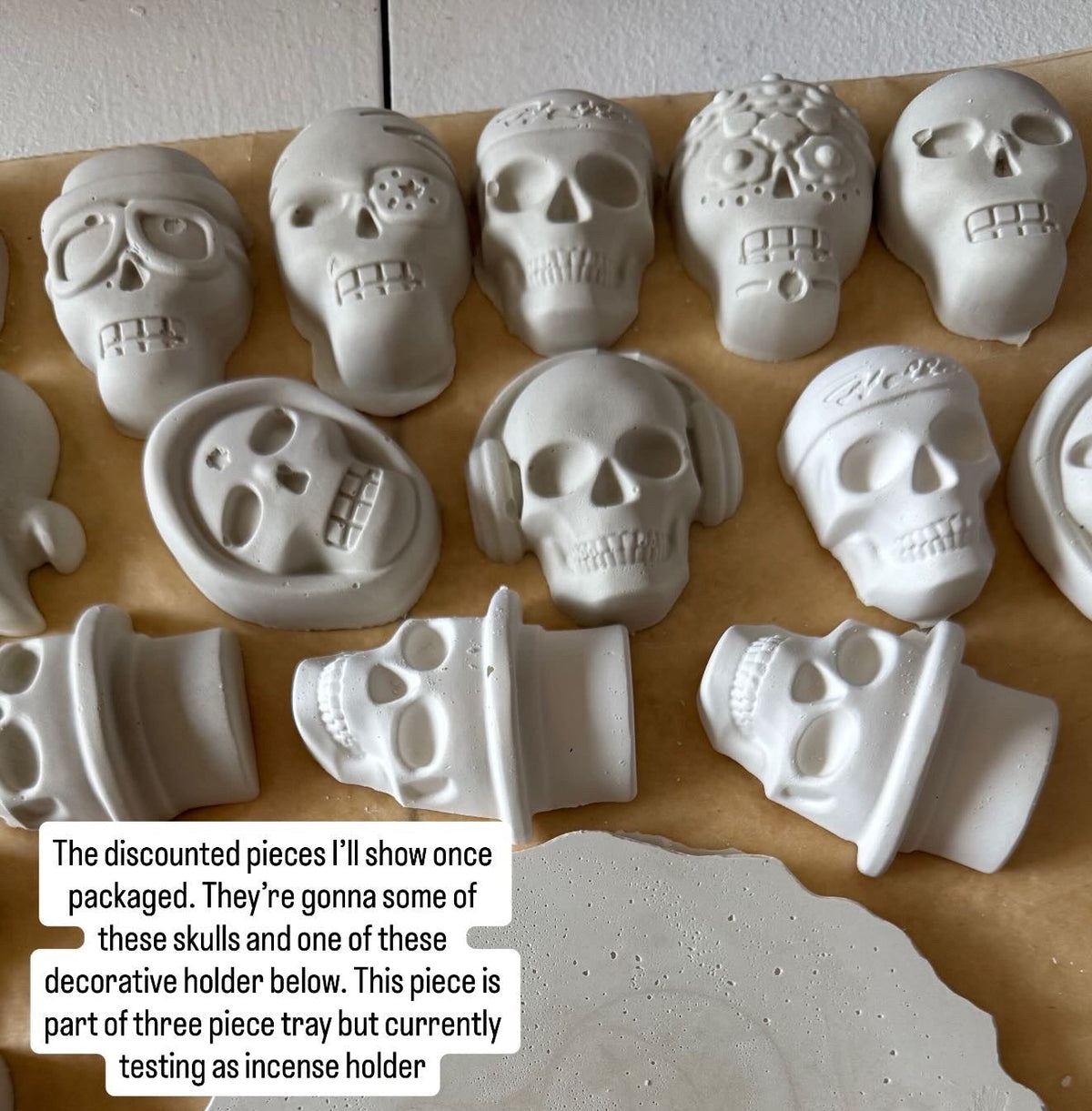 A collection of unpainted, light gray ceramic Day of the Dead decorative pieces, including sugar skulls, decorative holders, and a scalloped plate, arranged on brown kraft paper with a text overlay explaining the product selection.